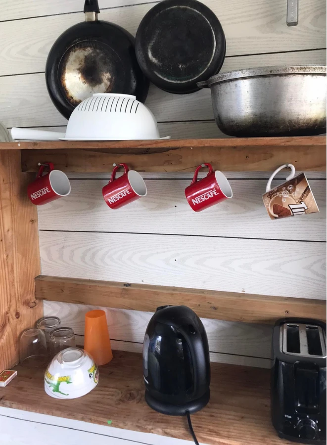 Le coin cuisine : poêles, marmite, passoire, tasses accrochées, bouilloire et grille-pain sur des étagères en bois